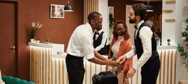 Happy african american tourists checking out from hotel after handing over room access card key to asian receptionist. Joyful guests tipping helpful bellboy carrying their luggage.