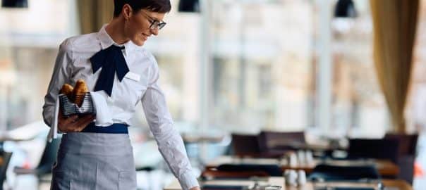 Happy hotel waitress serving breakfast at the tables in a restaurant.