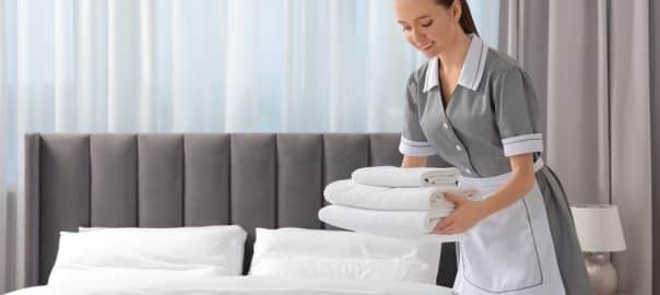 Young maid with stack of towels near bed in hotel room.