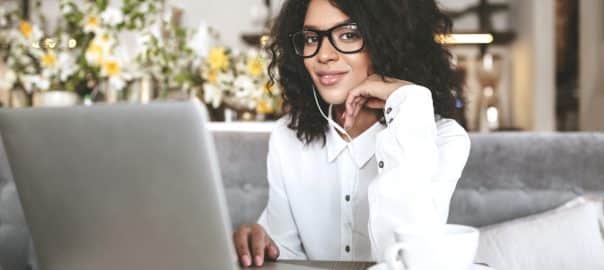 Nice African American girl in glasses sitting at restaurant with laptop and cup of coffee. Young lady with dark curly hair working on laptop in earphones.