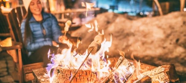 Winter holiday ski resort woman roasting marshmallows in BBQ firepit afterski fun leisure activity with friends. Couple grilling marshmallow stick in fire.