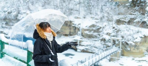 Happy Asian woman tourist in winter coat holding umbrella walking down small town street in snow day. Attractive girl enjoy and fun outdoor lifestyle travel countryside on winter holiday vacation.