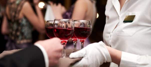 Close-up picture of waiter's hands wearing white gloves holding a tray with red wine, serving alcohol drinks. Catering service at special occasion, event. Hospitality industry concept.