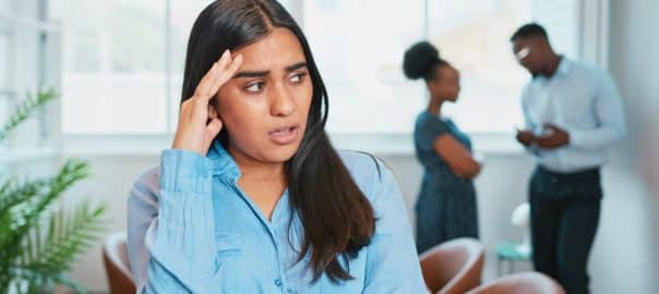 Young woman looks upset while colleagues talk politics at work.
