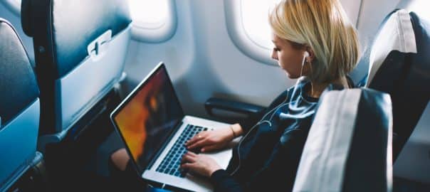 Attractive caucasian female passenger of airplane sitting in comfortable seat listening music in earphones while working at modern laptop computer with mock up area using wireless connection on board.
