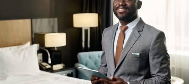 Waist up portrait of smiling African American man wearing suit as hotel manager standing in hotel room holding tablet and looking at camera copy space.