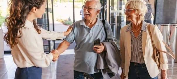 An elderly couple in casual travel attire stands in a hotel lobby, shaking hands with a friendly staff member, as they check in with their luggage beside them, ready to begin their stay