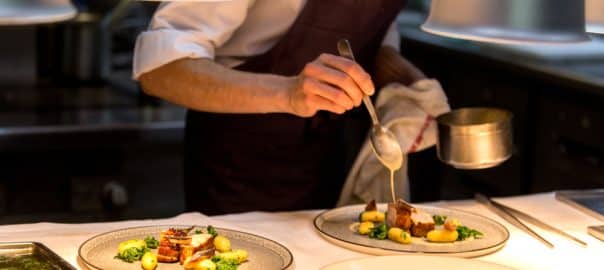 Chef preparing a plate made of meat and vegetables. The chef is pouring sauce on two plates.