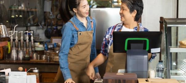 Young barista female hiring senior, elderly, 60s pensioner worker working in cafe bakery small business shop, two waitresses women standing behind coffee counter using technology cash register machine.