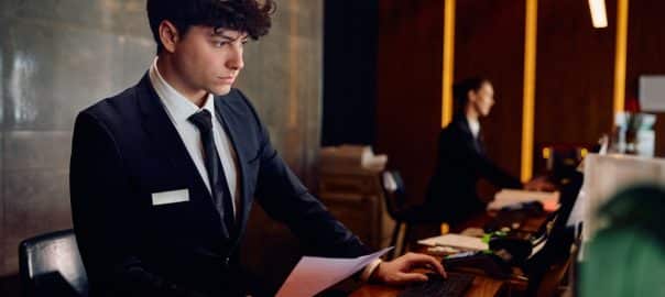 Young concierge working on a computer while checking documents at hotel reception desk.