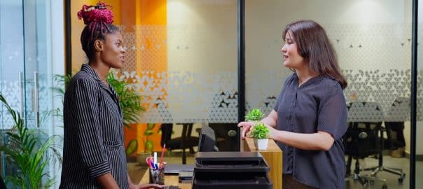 Happy young adult gen z confident receptionist woman standing reception desk table do welcome greeting to customer attend client talk typing registration data on computer indoor job office work place.