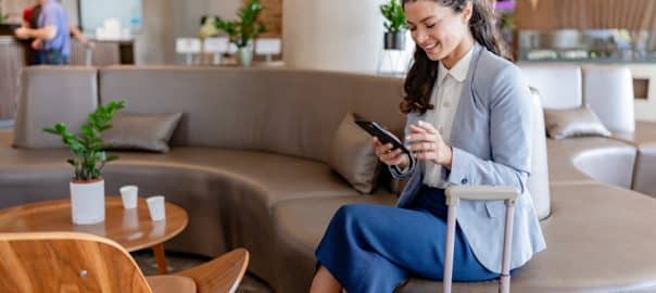 A smiling businesswoman sits in a modern hotel lobby, checking her phone while holding onto her suitcase. She appears relaxed and ready for her trip, surrounded by a comfortable lounge area.