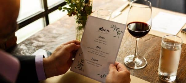 Man holding a menu at a restaurant table with a glass of red wine and water. Man reading menu with wine at the restaurant. Menu read by a man at restaurant's table with glass of wine and water.