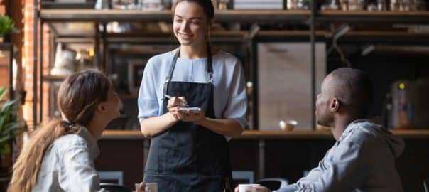 Smiling millennial waitress with notebook taking order from young multiracial client couple, diverse friends relax hang out in cafe or restaurant speak with staff enjoying good service and atmosphere.