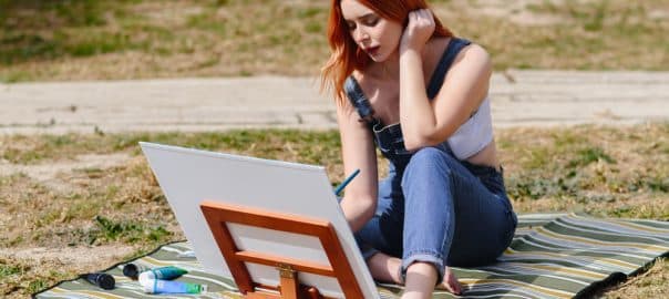 Young caucasian redhead female artist paints a canvas on a small easel with a brush in the park outdoors.