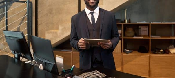 Businessman in formal attire standing in modern office environment holding a tablet. Smiling while looking at camera in an organized office setting.