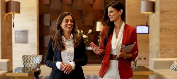 Two businesswomen walking and discussing in a modern hotel lobby, collaborating and smiling during a professional meeting.