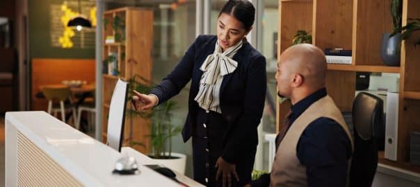 Man, woman and concierge with mentor at hotel with computer, point and discussion for booking info. People, training and receptionist with listening with pc for hospitality with coach at lodge.