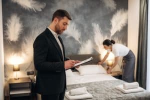 Male,Hotel,Manager,Reviews,Notes,In,A,Modern,Hotel,Room Male hotel manager reviews notes in a modern hotel room as a female staff member adjusts the bedding, showcasing a stylish interior design.